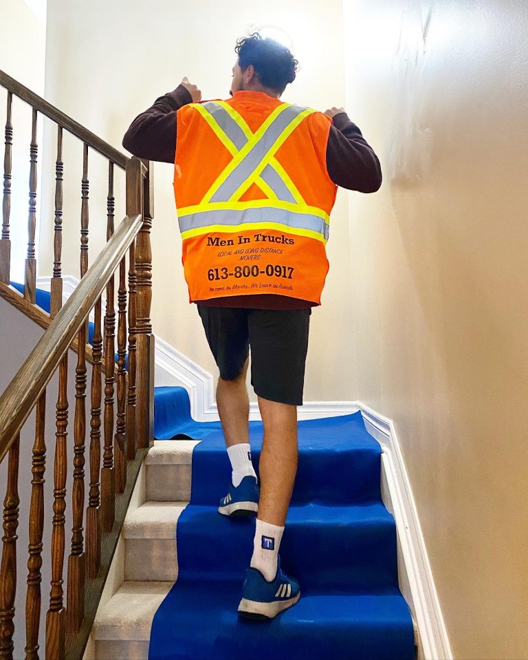 Mover in an orange vest climbing a staircase covered by a blue floor runner.